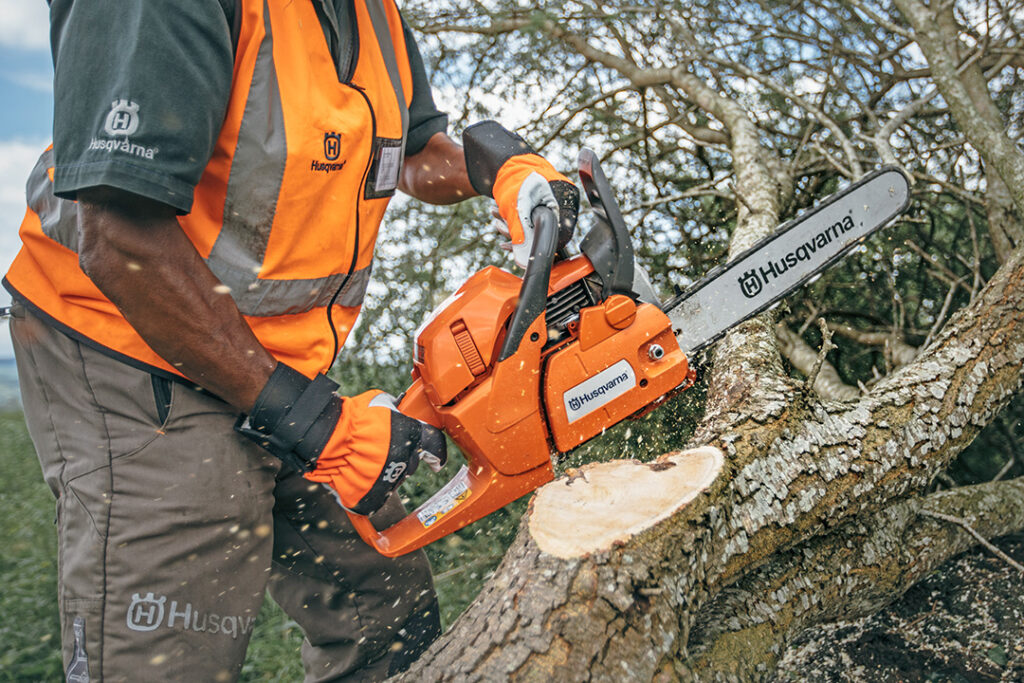 A man holds a Husqvarna chainsaw while cutting through a branch on a felled tree.