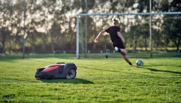 A Husqvarna Automower cutting the grass on a soccer field while a player kicks a ball at the goals in the background.