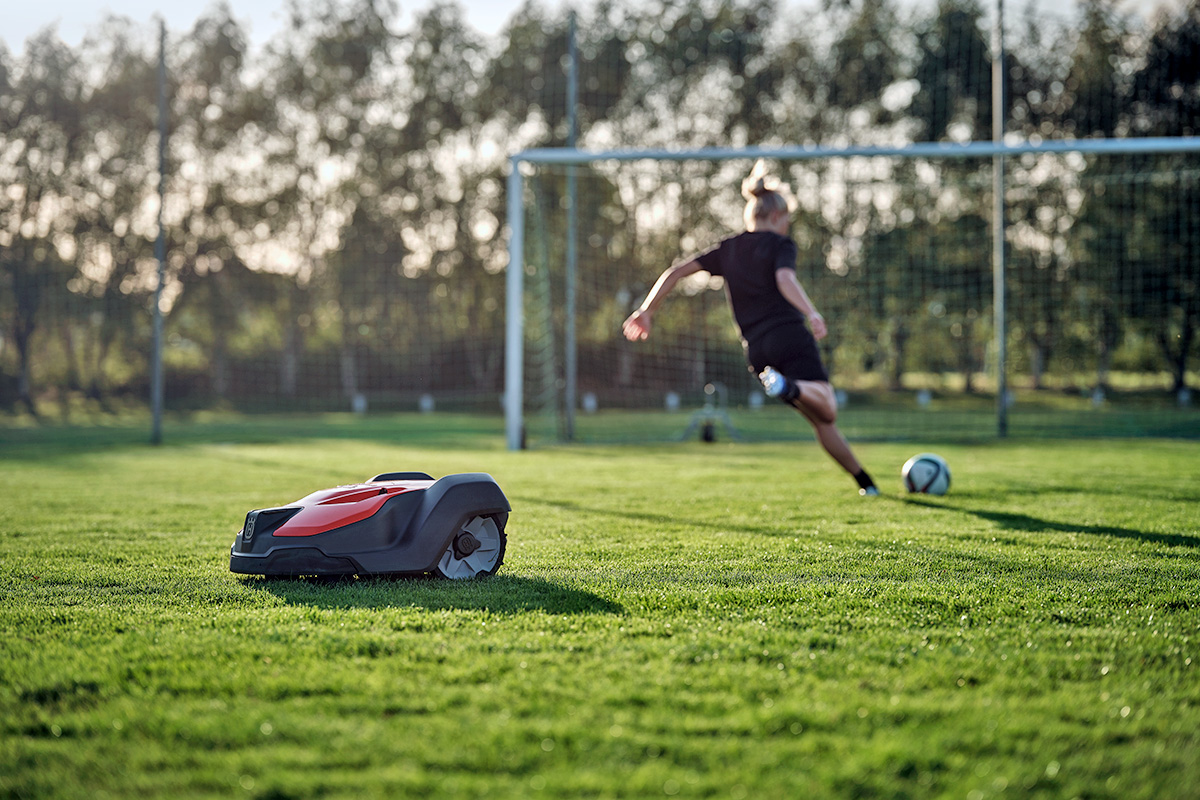 A Husqvarna Automower cutting the grass on a soccer field while a player kicks a ball at the goals in the background.