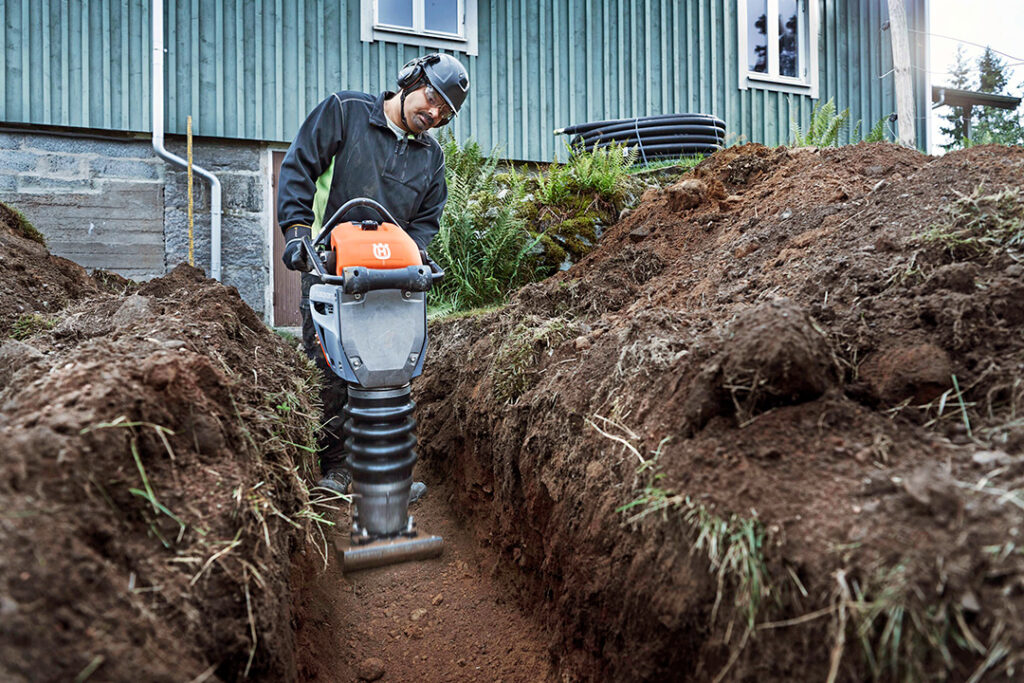 A man working with a Husqvarna tamping rammer in a foundation ditch.