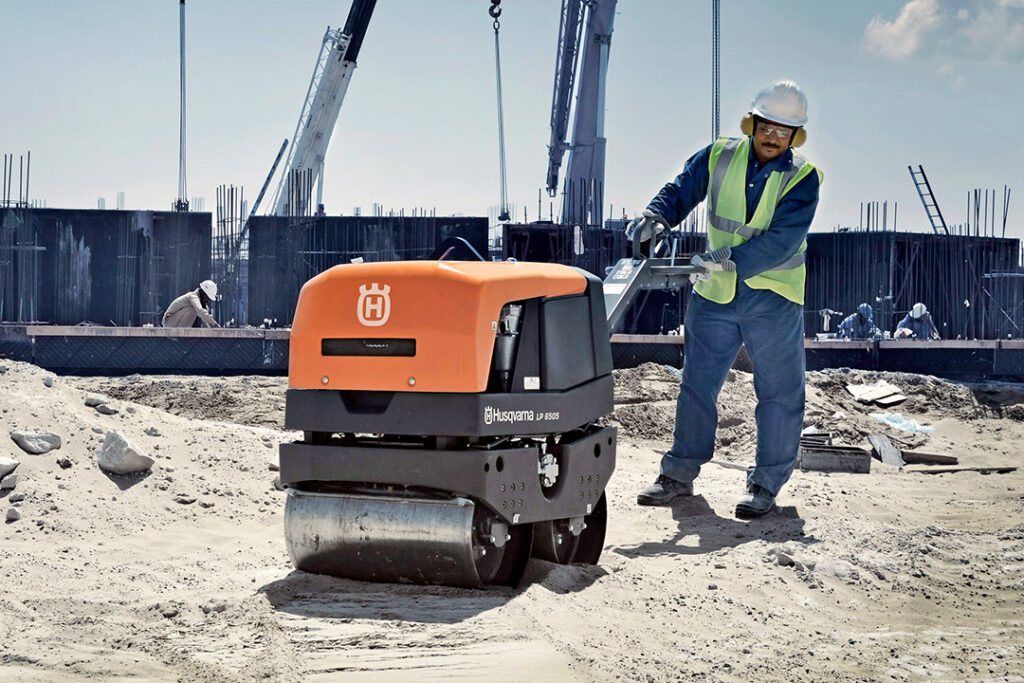 A construction worker using a Husqvarna roller compactor on a sandy surface.