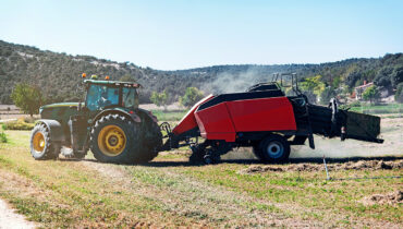 A farmers in a green tractor pulling a red trailer through a dusty field.