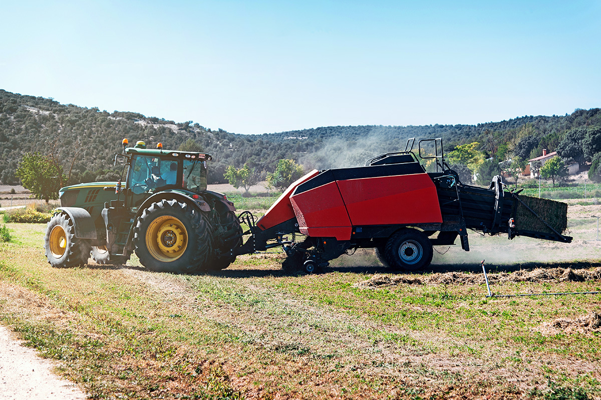 A farmers in a green tractor pulling a red trailer through a dusty field.