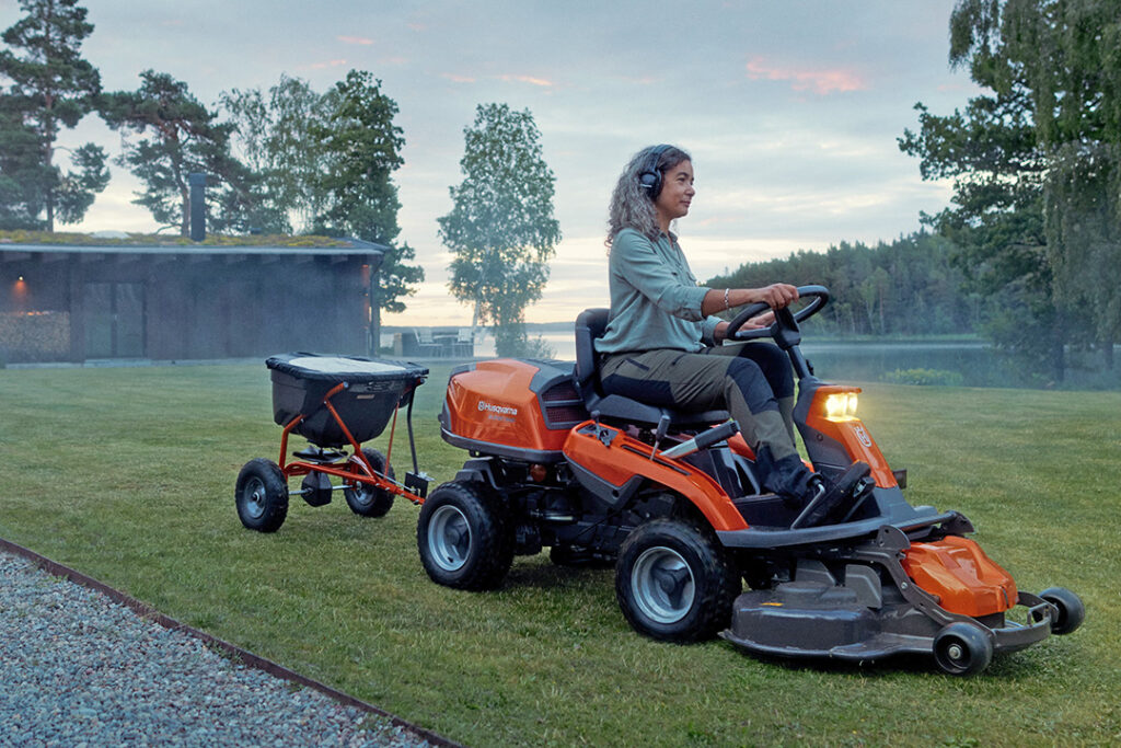 A woman driving a Husqvarna R316T rider with a tow spreader attached to the back.
