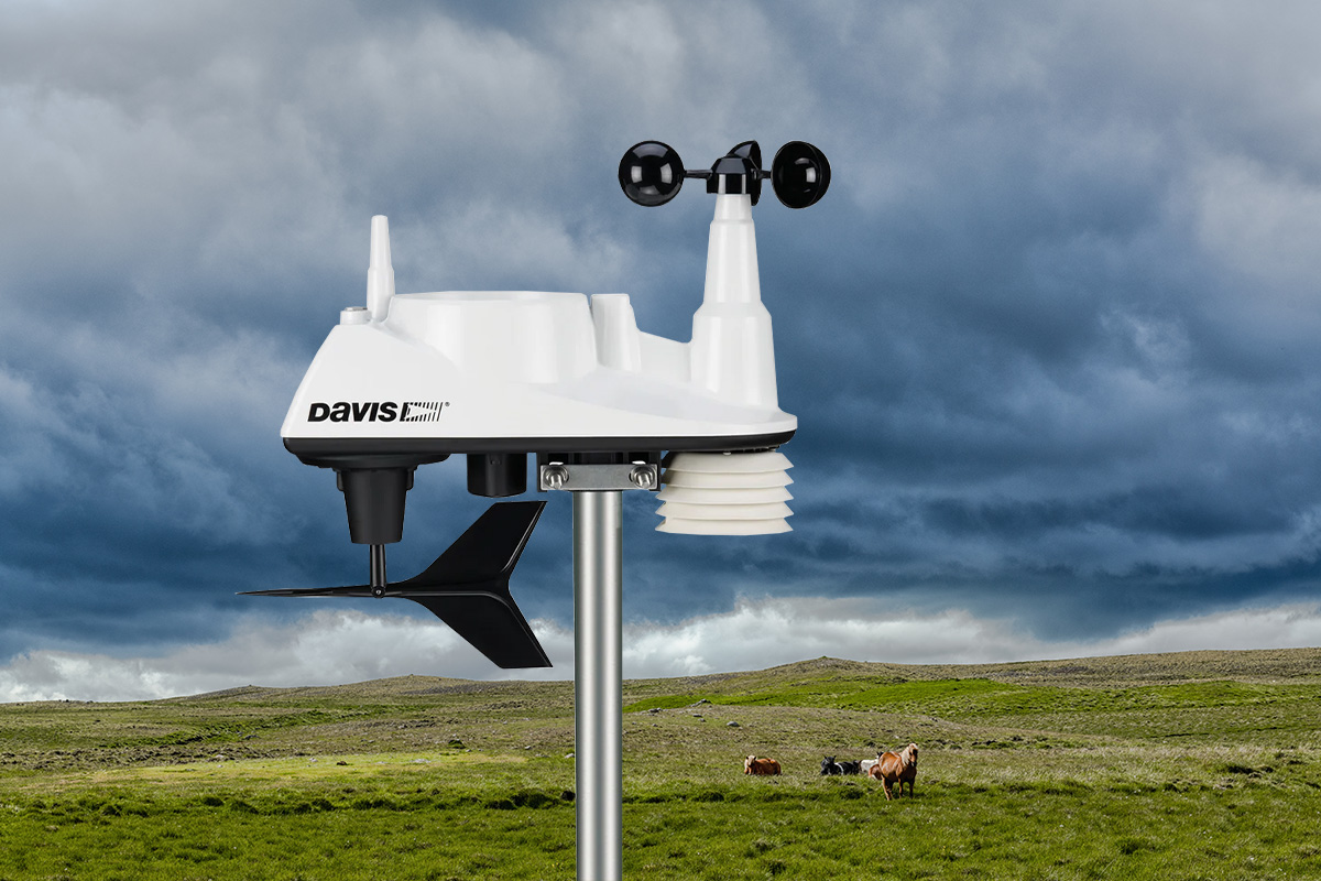 A Davis weather station with a moody sky and farm in the background